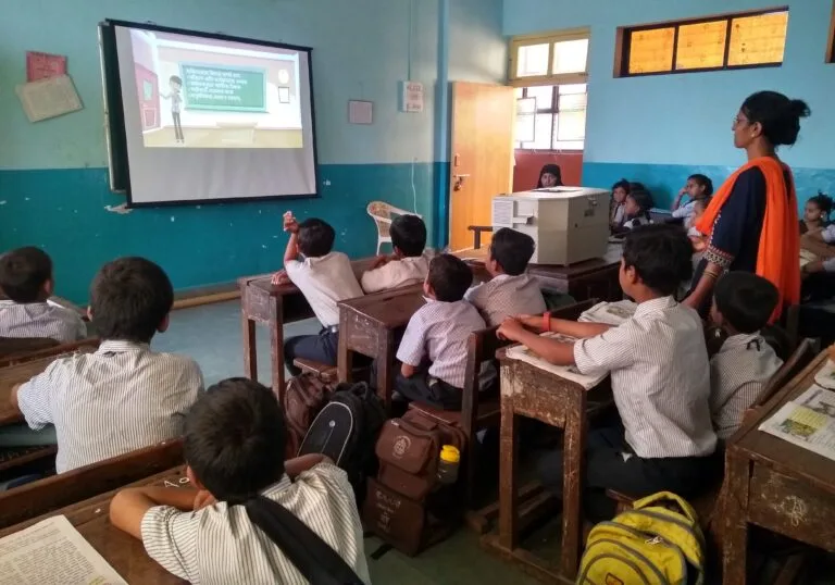 EdConnect-2-e1570793855828 - Best Study Abroad Consultants in India Classroom of young students in school uniforms attentively watching an educational video projected on a screen, with a teacher standing nearby guiding the lesson.